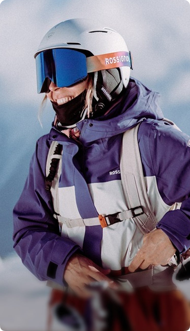 Person wearing a white helmet, blue mirrored ski goggles, and a purple‑and‑white ski outfit, standing outdoors in a snowy environment.