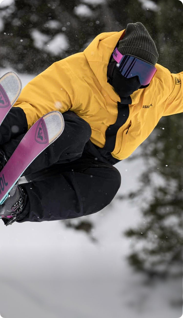 Sciatore che esegue una figura freestyle nella neve, vestito con una giacca gialla Desafio 2L e una maschera Otava viola.