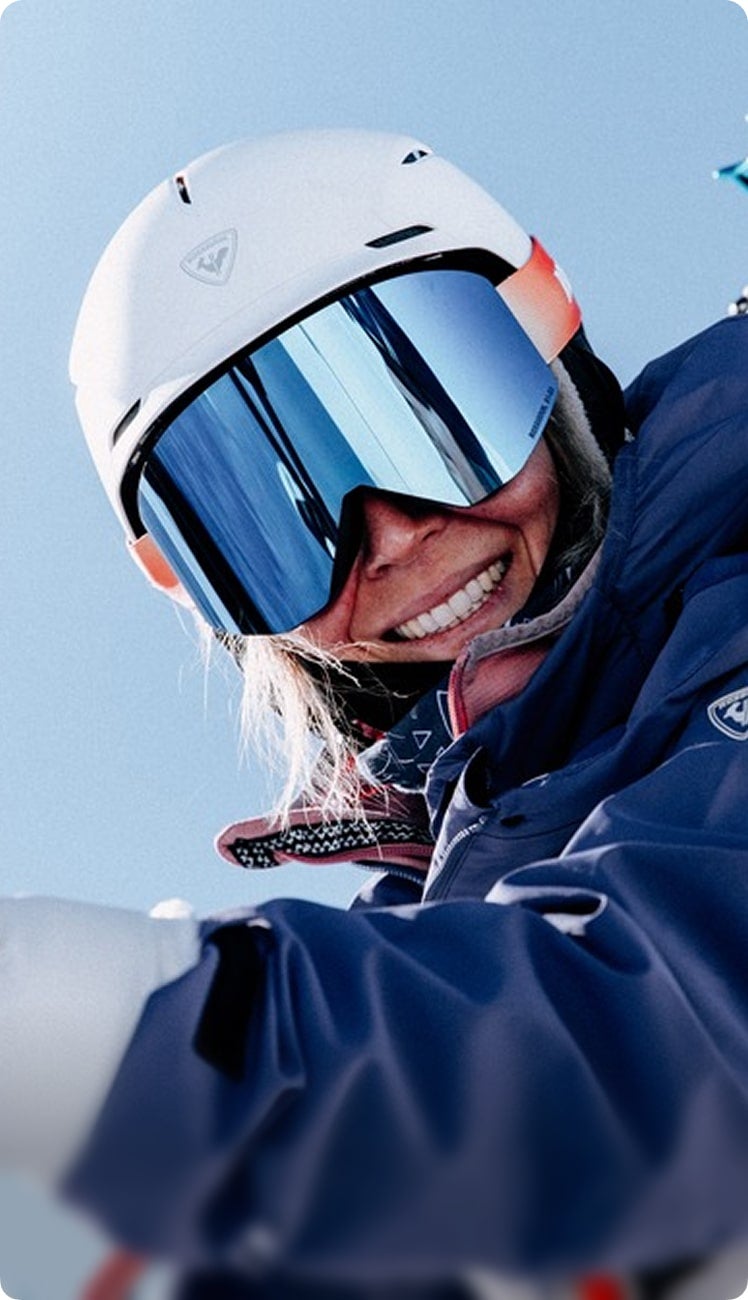 Skier wearing a white helmet and blue mirrored ski goggles, smiling while dressed in navy ski outerwear in a snowy mountain setting.