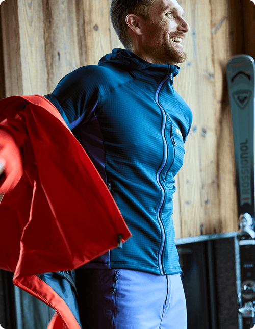 Mann steht und trägt ein blaues Midlayer, während er eine rote Rochrun-Skijacke anzieht.