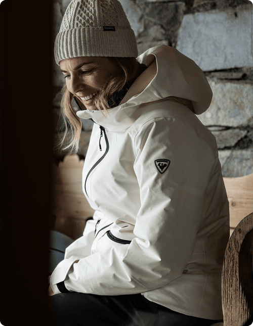 Skifahrerin sitzt und trägt eine weiße Rochrun-Skijacke mit einer beigen Opal-Mütze.