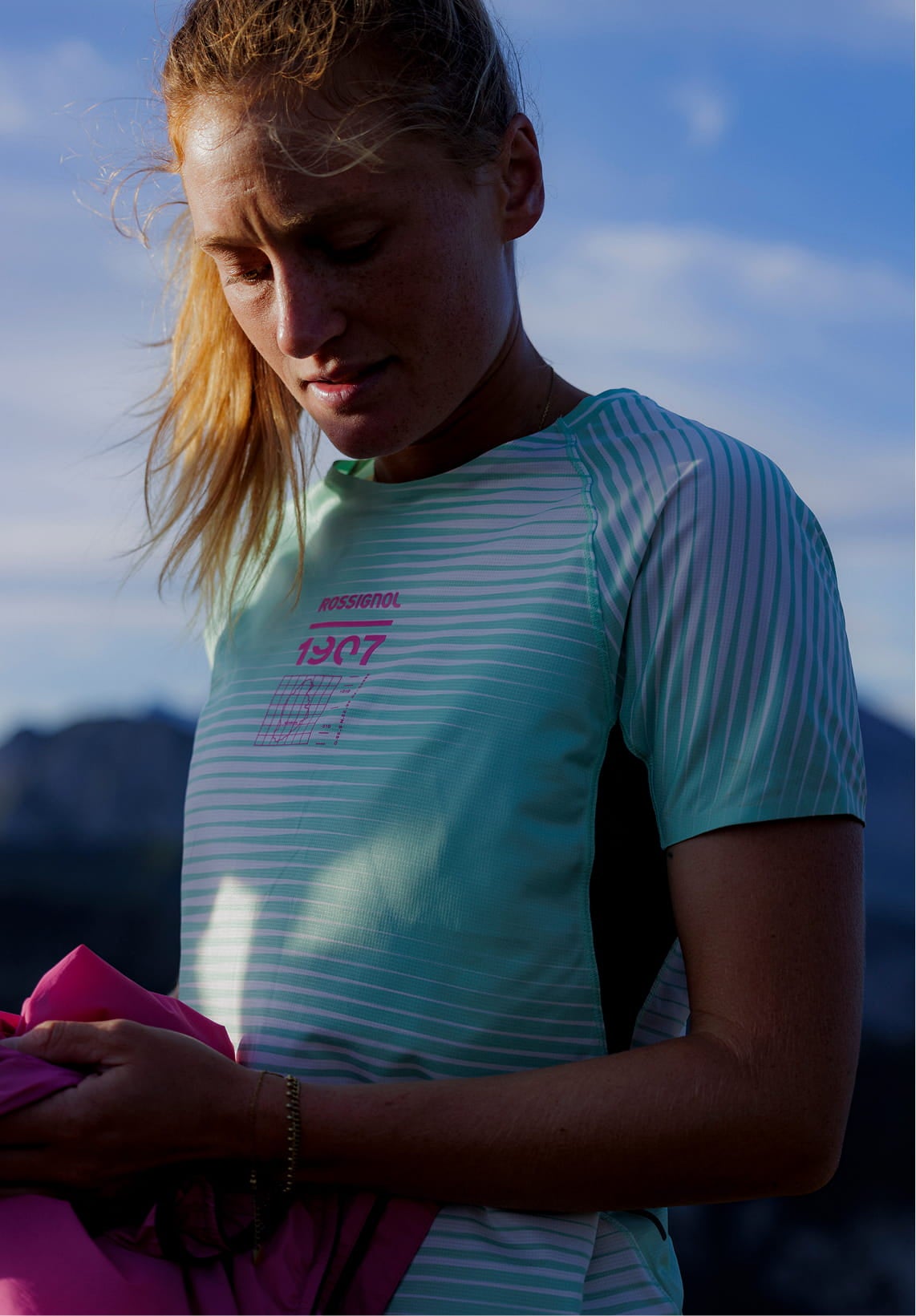 Person standing outdoors on a mountain trail, holding a pink jacket and wearing a light green technical T‑shirt with graphic patterns.