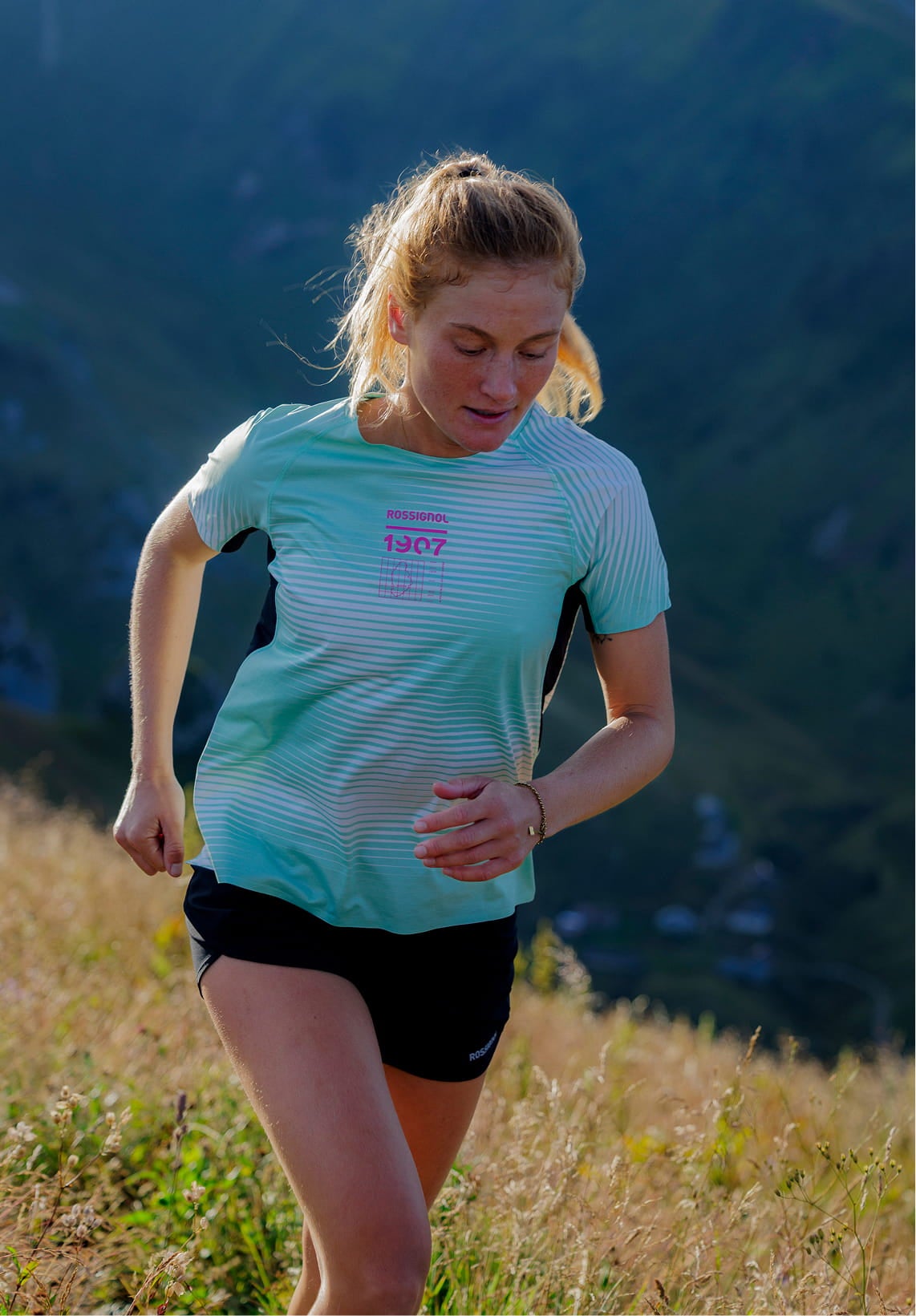 Person running through tall grass on a mountain trail, wearing a light green patterned technical T‑shirt and dark shorts.