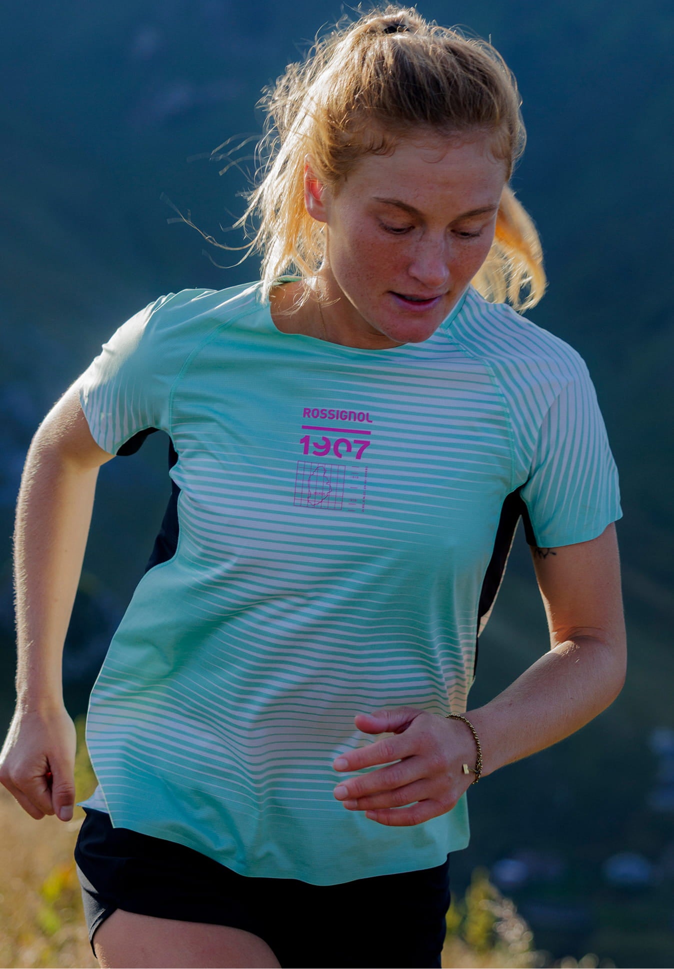 Person running through tall grass on a mountain trail, wearing a light green patterned technical T‑shirt and dark shorts.