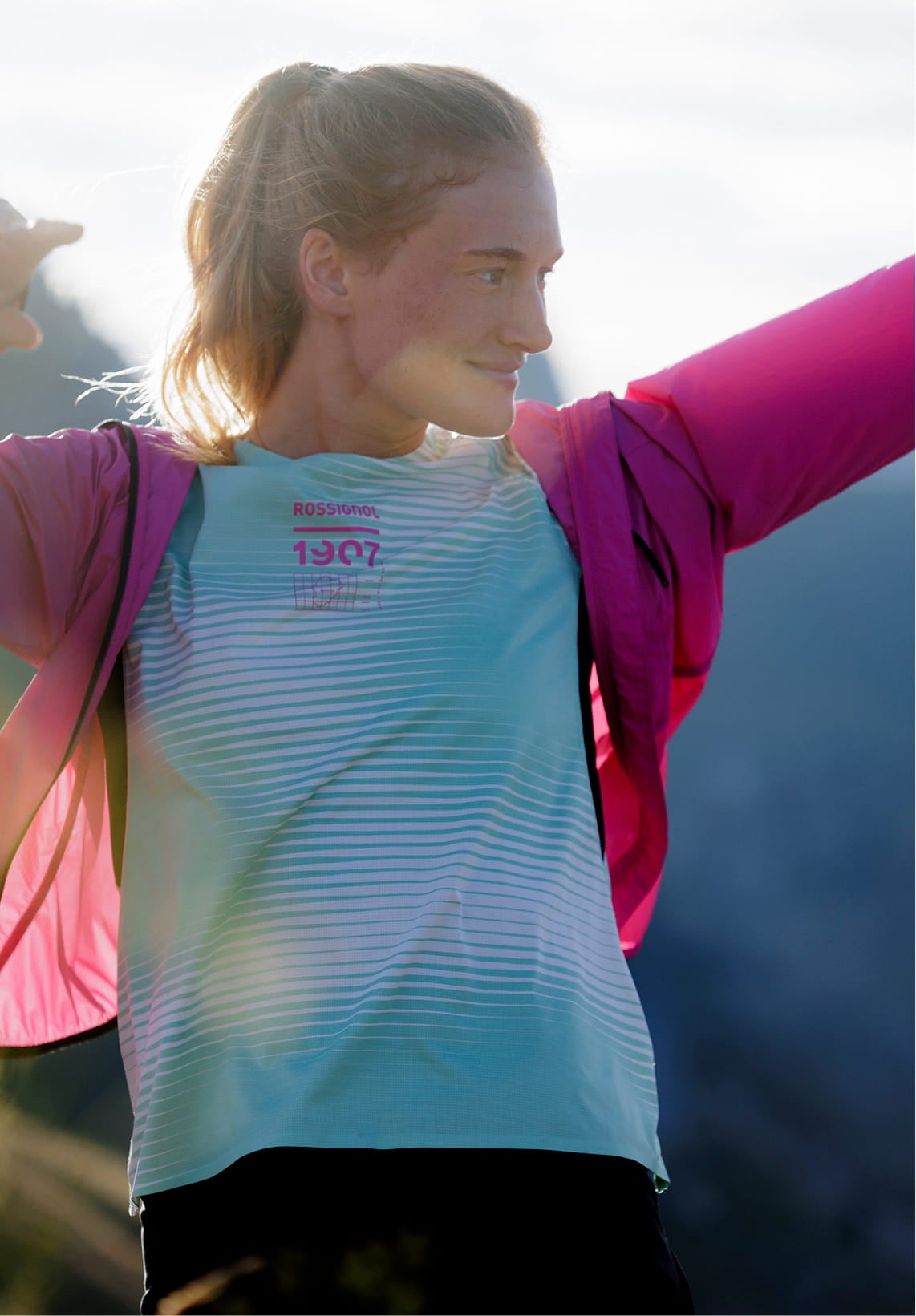 Person on a mountain trail raising one arm while holding a pink jacket, wearing a light green technical T-shirt with graphic patterns.