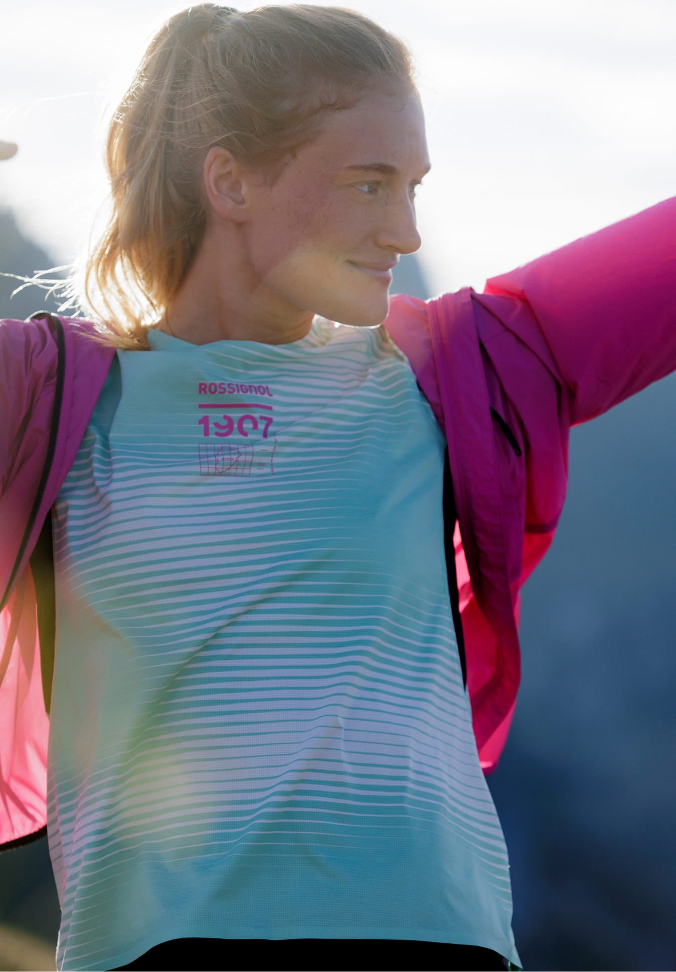 Person on a mountain trail raising one arm while holding a pink jacket, wearing a light green technical T-shirt with graphic patterns.