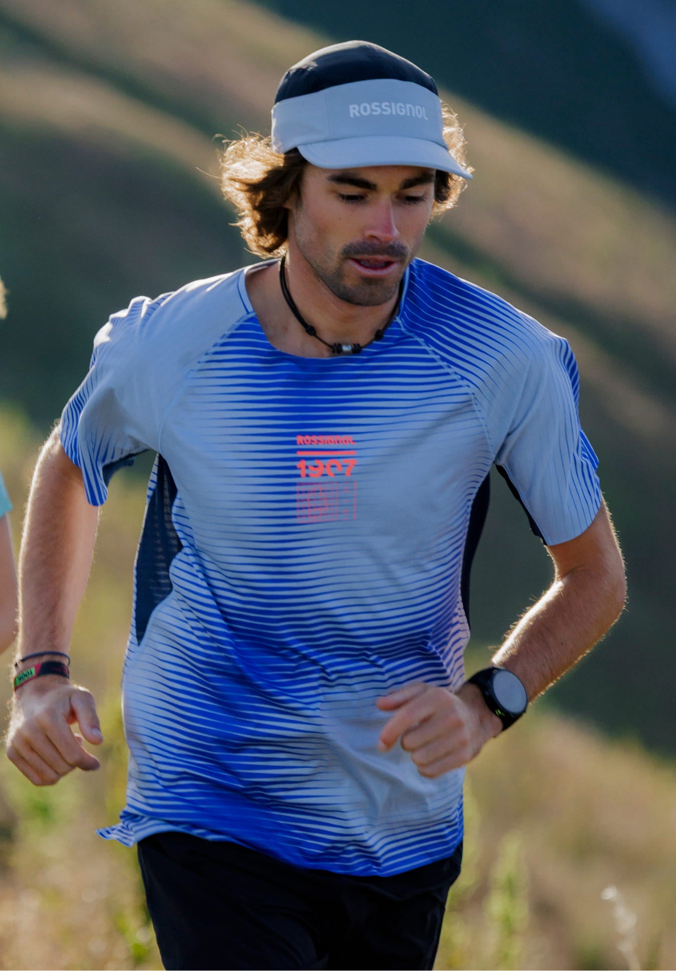 Person running on a mountain trail wearing a blue Rossignol technical T-shirt with linear patterns.