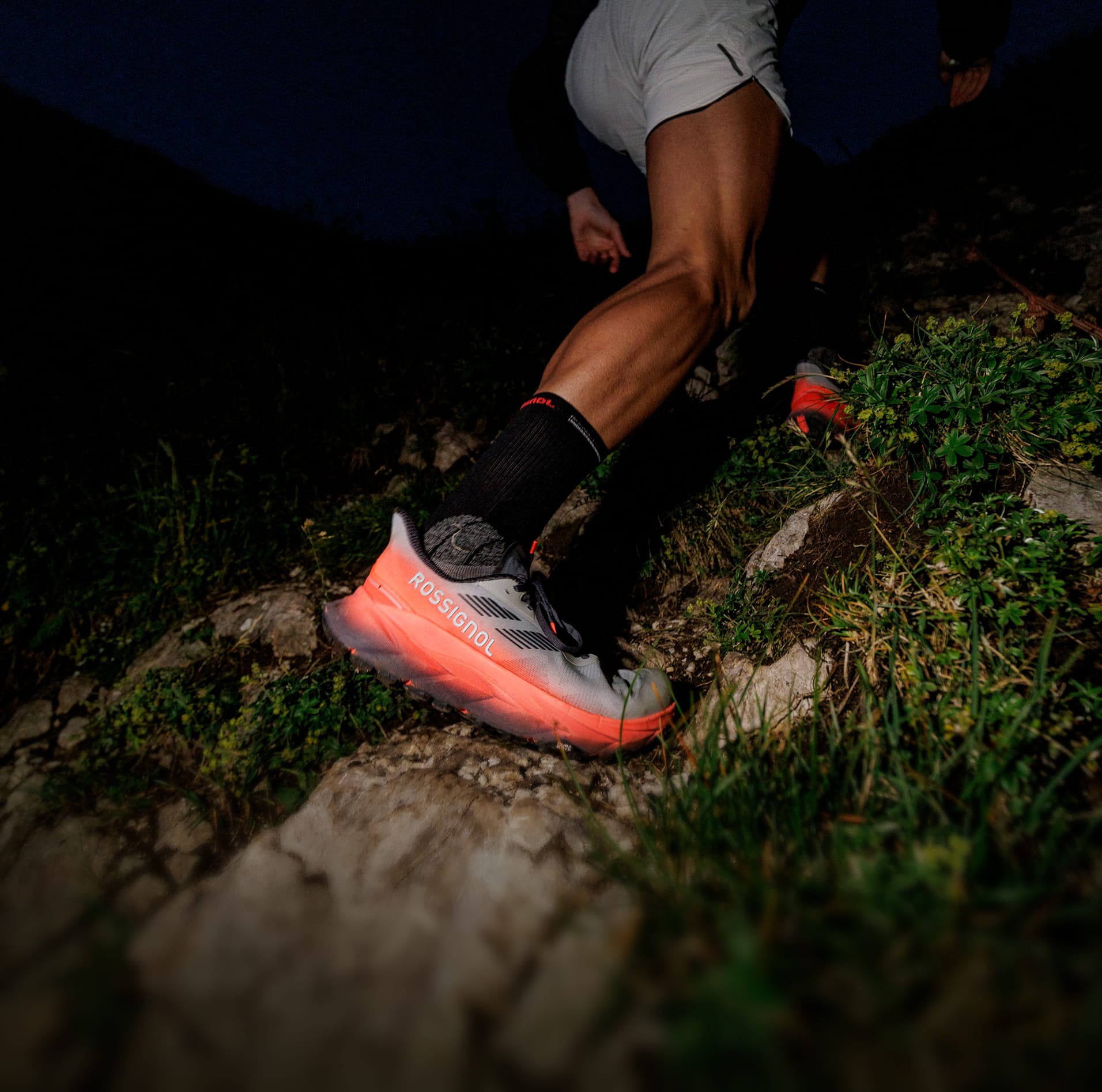 Close-up of a person running uphill on a rocky trail, wearing orange and black trail shoes, with a dark background indicating low-light conditions.
