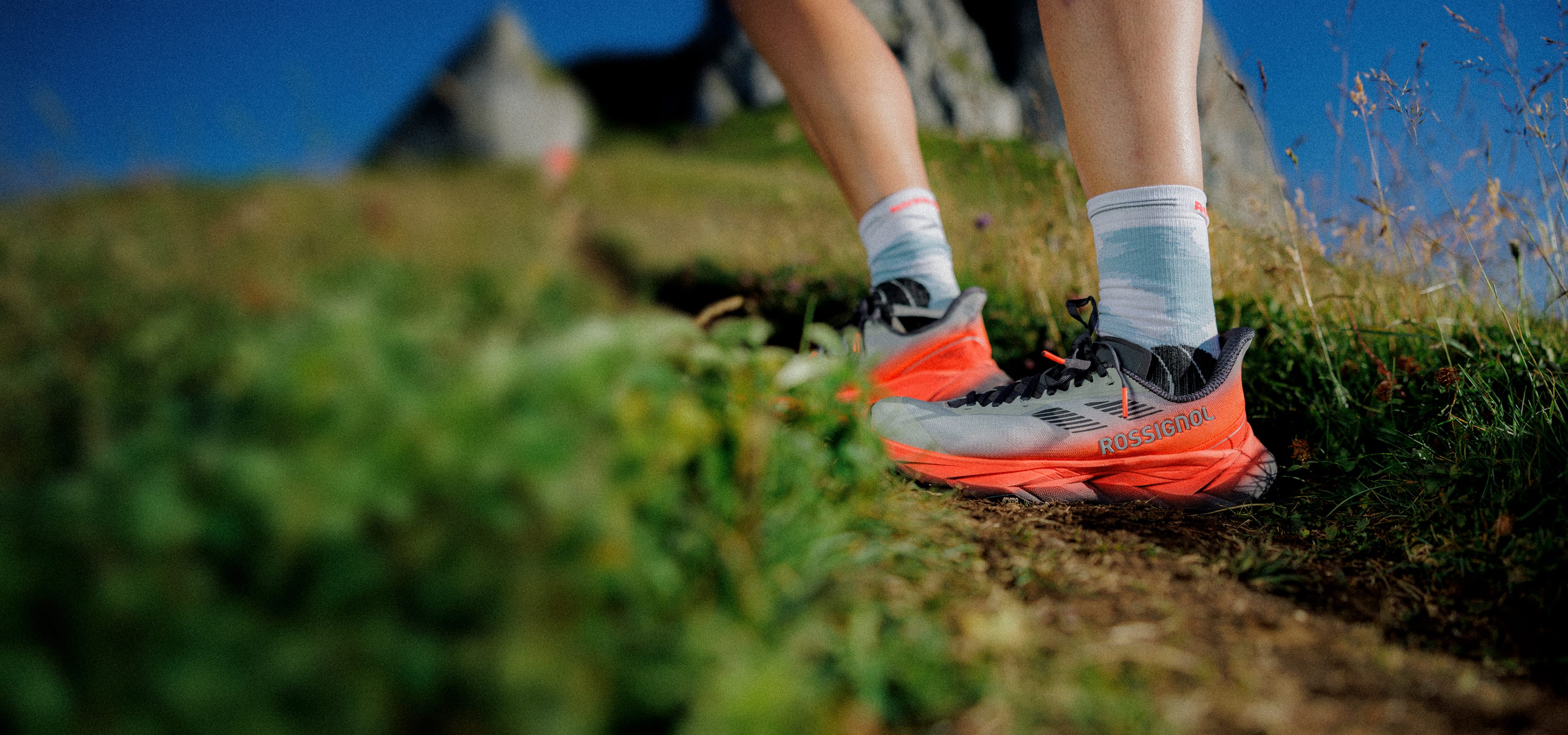 Close-up of a person wearing Vercors trail running shoes on a narrow dirt path, surrounded by grass and rocks.