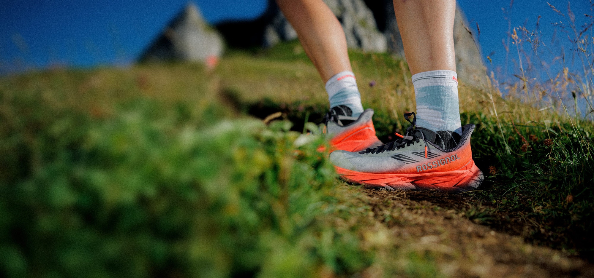 Close-up of a person wearing Vercors trail running shoes on a narrow dirt path, surrounded by grass and rocks.