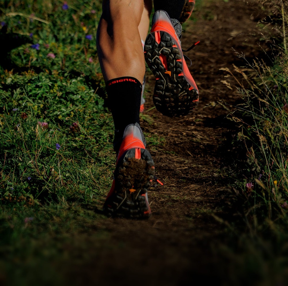 Close-up of trail running shoes on a narrow dirt and gravel path, with low vegetation lit by warm light.
