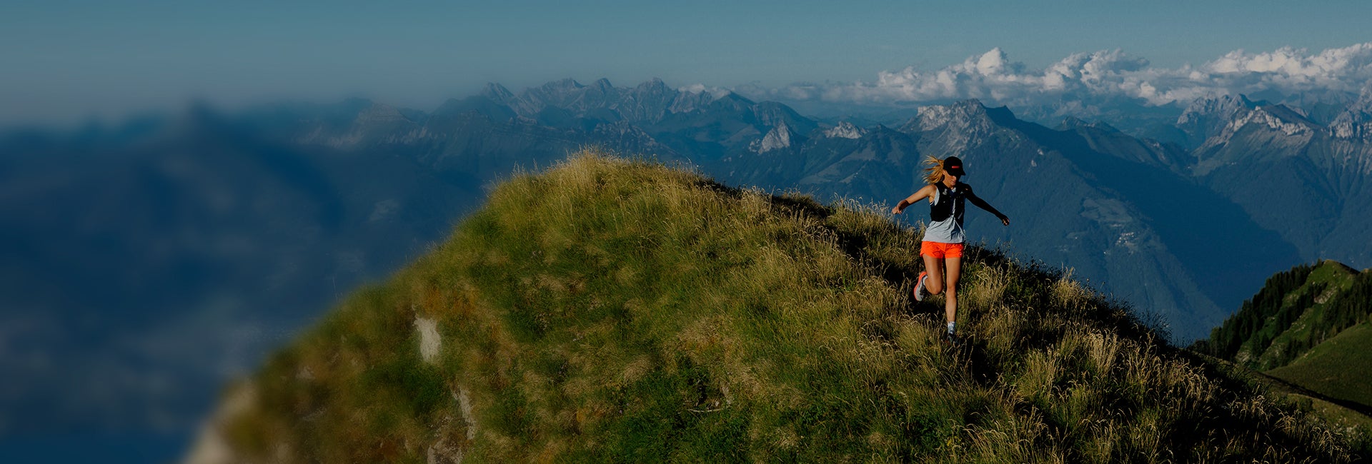 Une personne court sur une crête herbeuse en montagne, avec une vaste chaîne de montagnes et un ciel bleu en arrière-plan.