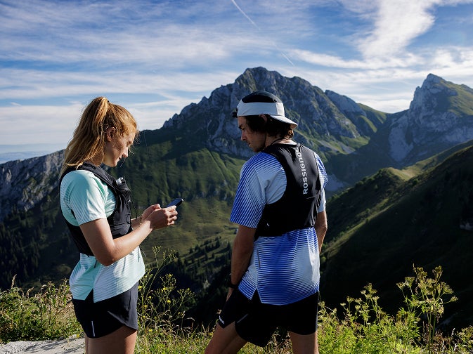 Deux personnes font une pause sur un sentier de montagne et consultent un téléphone, avec des sommets en arrière-plan.