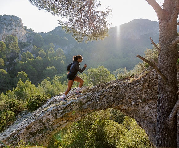 Une personne court en montée sur un chemin rocailleux en montagne, entouré d’arbres et de végétation.