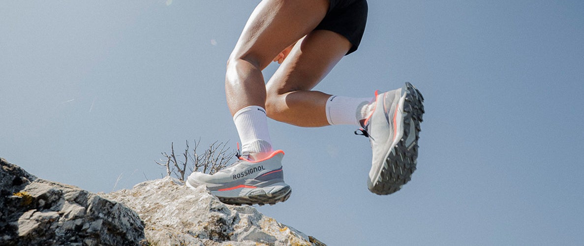 Gros plan sur les jambes d’une personne courant sur un terrain rocheux en montagne, avec des chaussures de trail en appui sur les pierres.