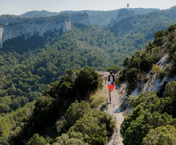 Une personne court sur un sentier étroit de montagne, bordé de végétation, avec un vaste paysage naturel en arrière-plan.