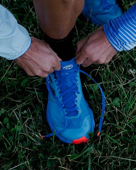 Gros plan sur des mains serrant les lacets d’une chaussure de trail sur l’herbe.