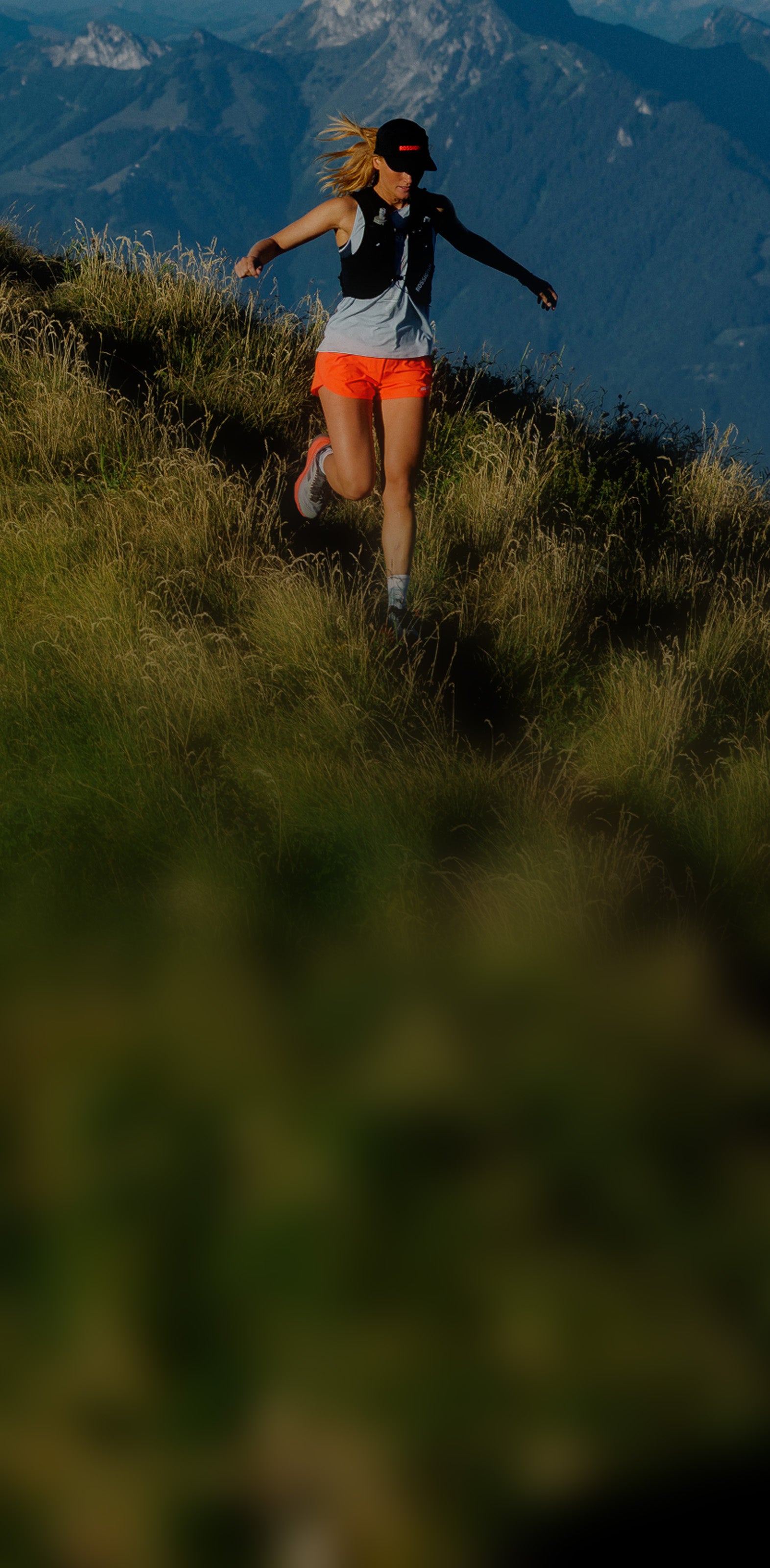 A person running along a grassy mountain ridge, with a wide mountain range and blue sky in the background.