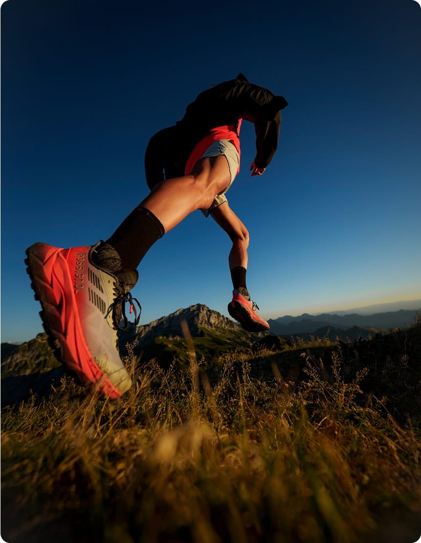 Red Vercors trail running shoe in motion during a dynamic uphill run on a grassy mountain path.