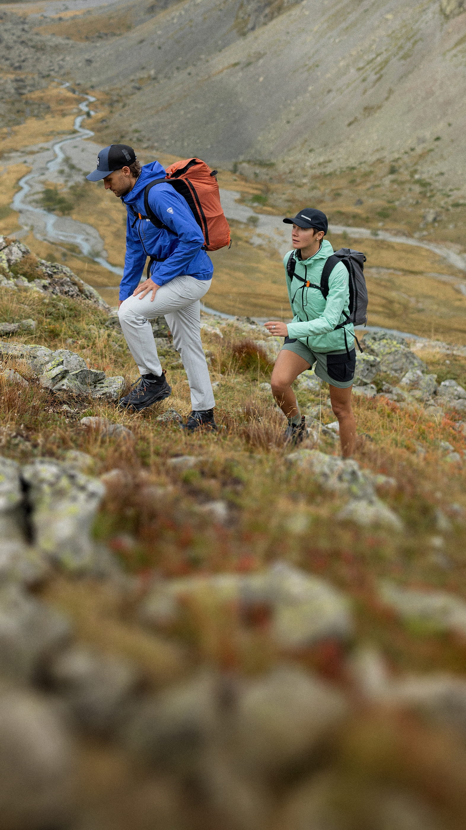 Two hikers climbing uphill on mountainous terrain, wearing hiking jackets and backpacks, with a wide valley landscape in the background.
