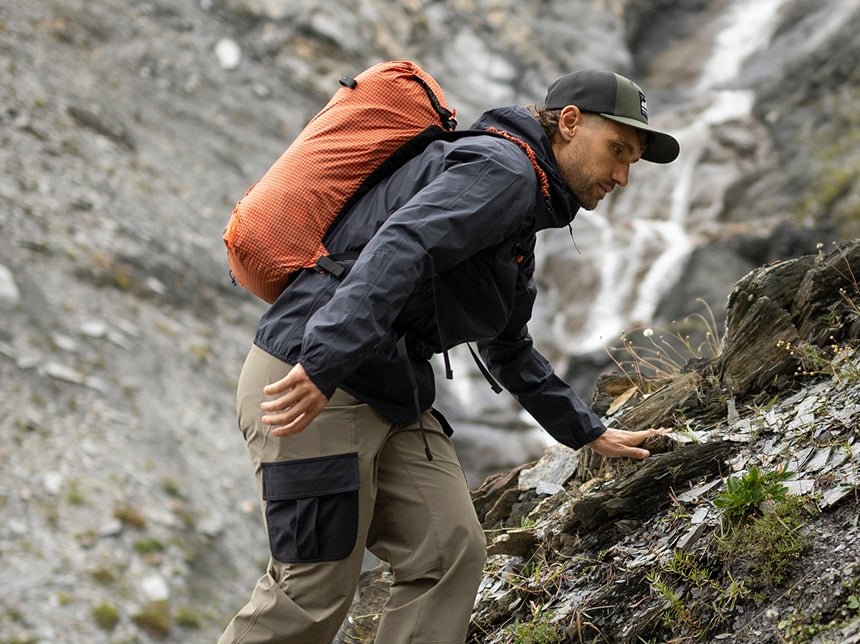  Une personne équipée d’un sac à dos progresse sur un sentier rocheux humide en montagne, à proximité d’une cascade.