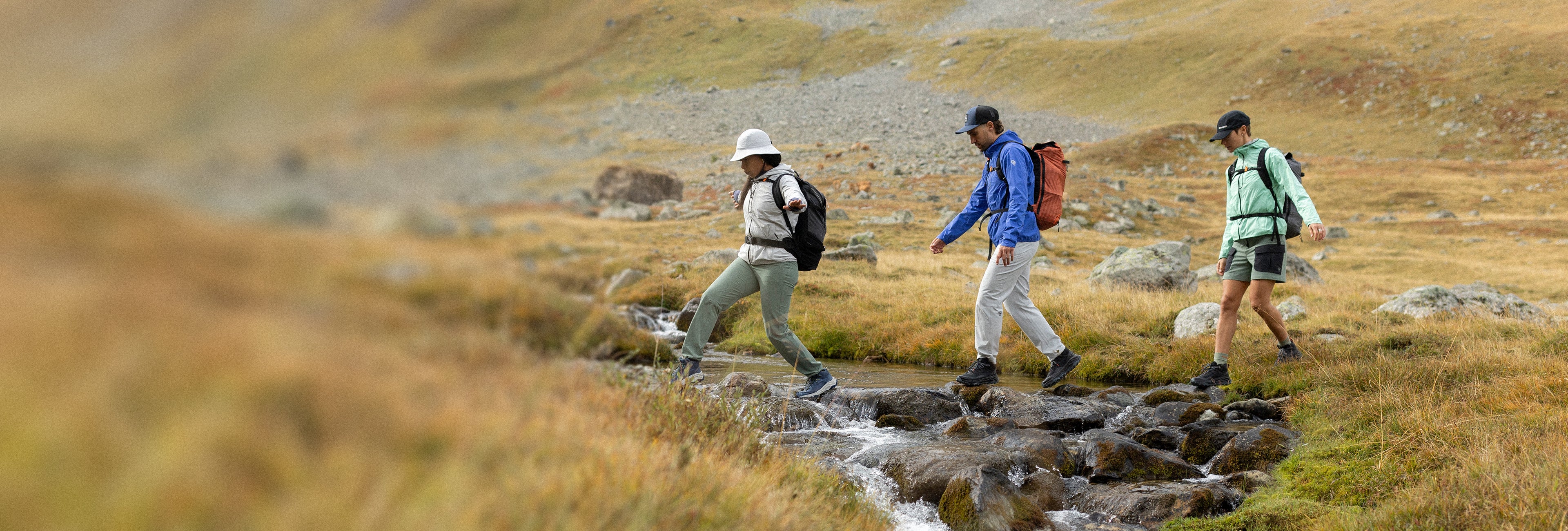 Trois personnes équipées de sacs à dos et de vêtements de randonnée traversent un petit ruisseau à pied sur des pierres, dans un paysage de montagne herbeux et rocailleux.