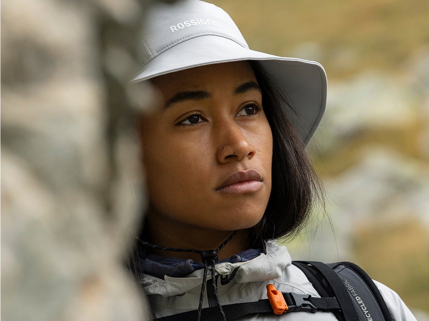 Portrait rapproché d’une personne portant un chapeau et une veste de randonnée, avec un paysage montagneux flou en arrière-plan. 