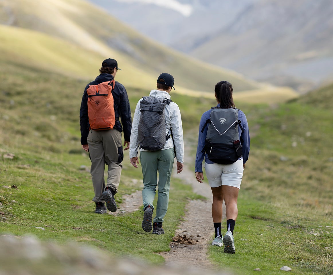 Trois personnes équipées de sacs à dos marchent sur un sentier de montagne herbeux, vues de dos, dans un paysage alpin vallonné. 