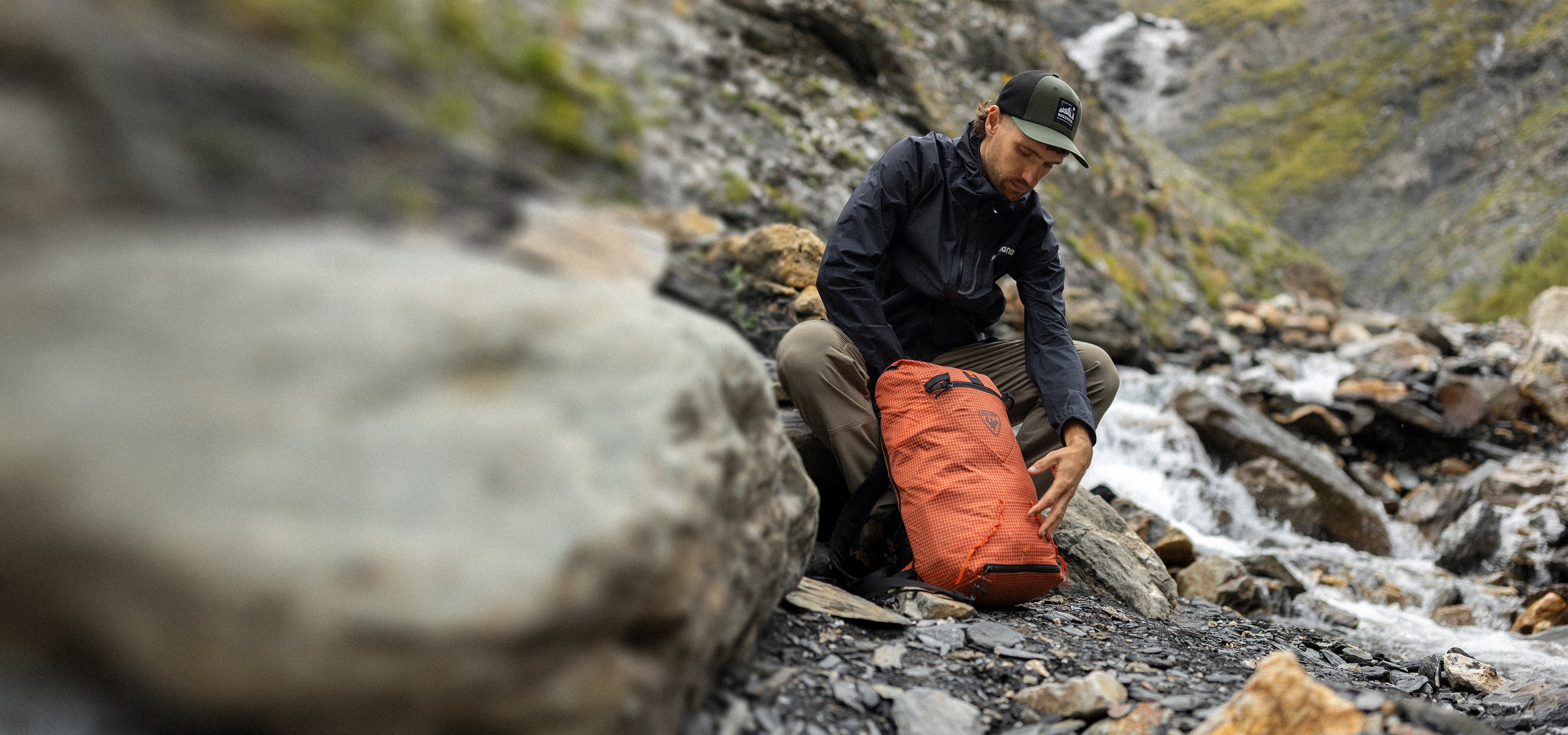  Une personne en tenue de randonnée est accroupie près d’un ruisseau de montagne, en train d’ajuster ou de vérifier un sac à dos orange posé sur des rochers. 