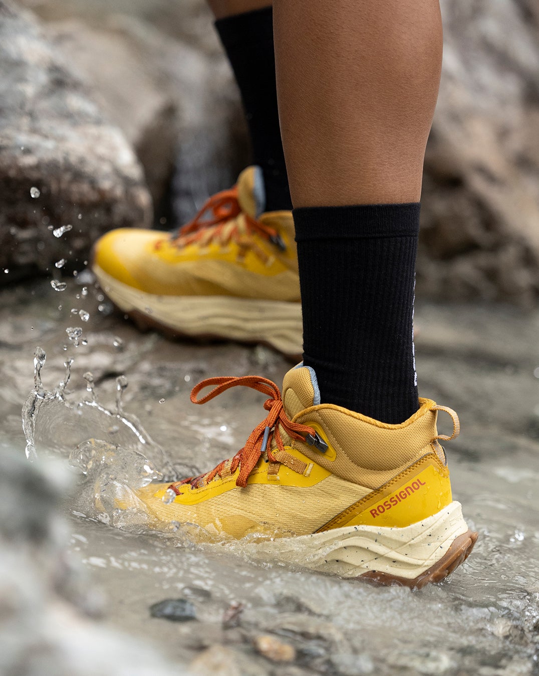 Gros plan sur une chaussure de randonnée Altiridge Mid de Rossignol, de couleur jaune, marchant dans un ruisseau avec de l’eau éclaboussée autour du pied. 