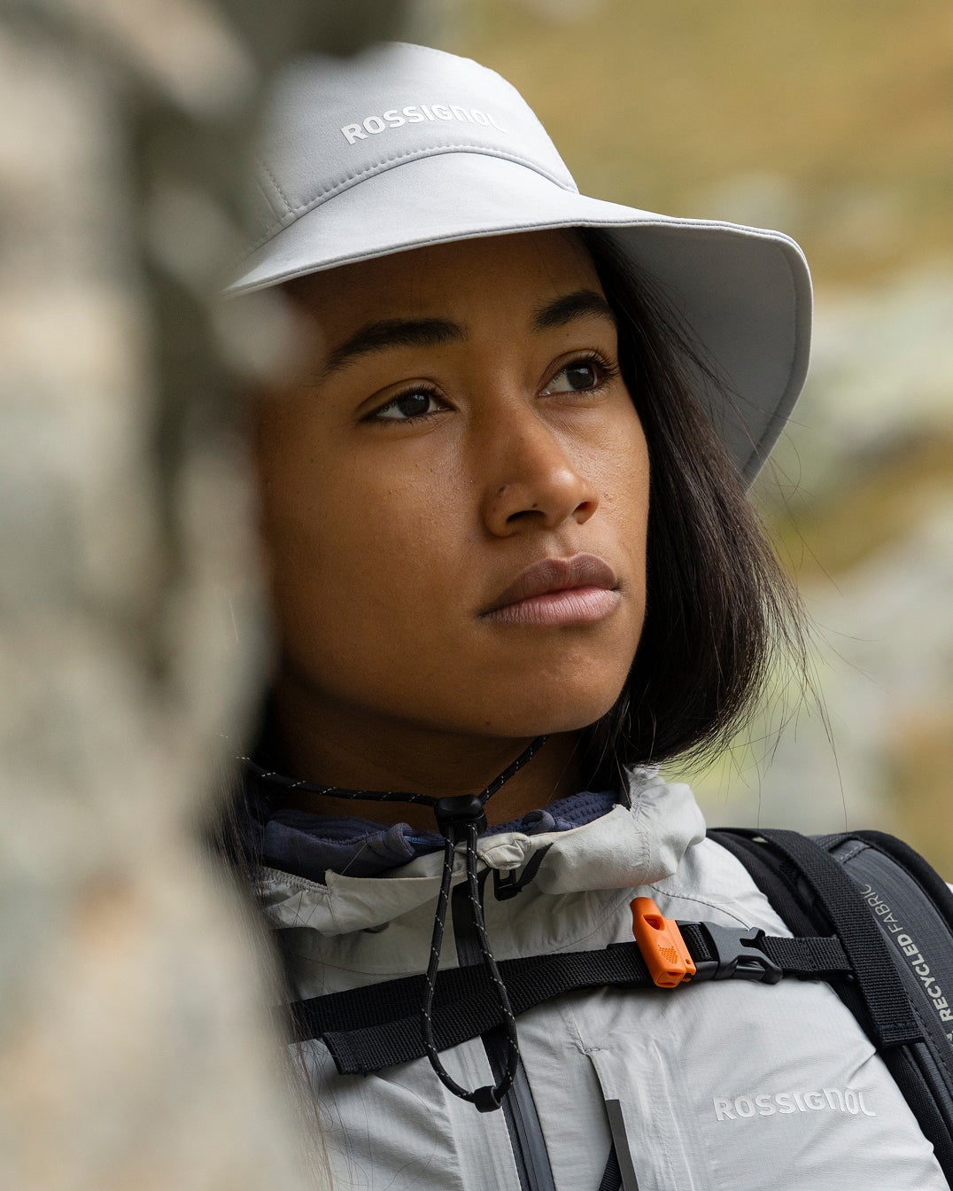 Close-up portrait of a person wearing a hiking hat and jacket, with a blurred mountain landscape in the background.