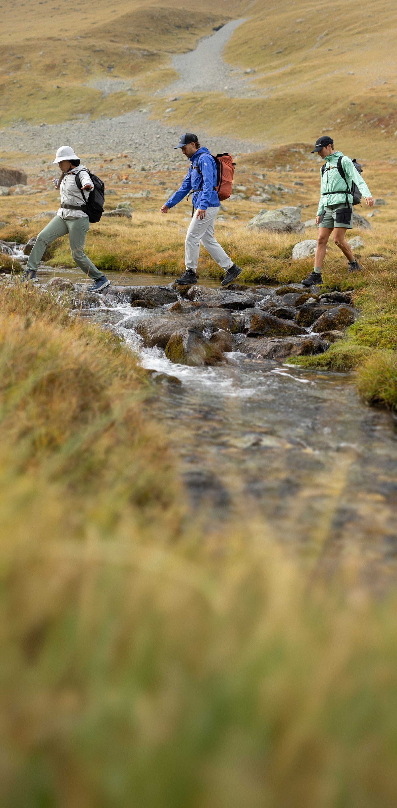 Trois personnes équipées de sacs à dos et de vêtements de randonnée traversent un petit ruisseau à pied sur des pierres, dans un paysage de montagne herbeux et rocailleux. 