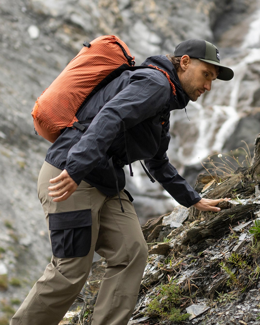  Une personne équipée d’un sac à dos progresse sur un sentier rocheux humide en montagne, à proximité d’une cascade. 