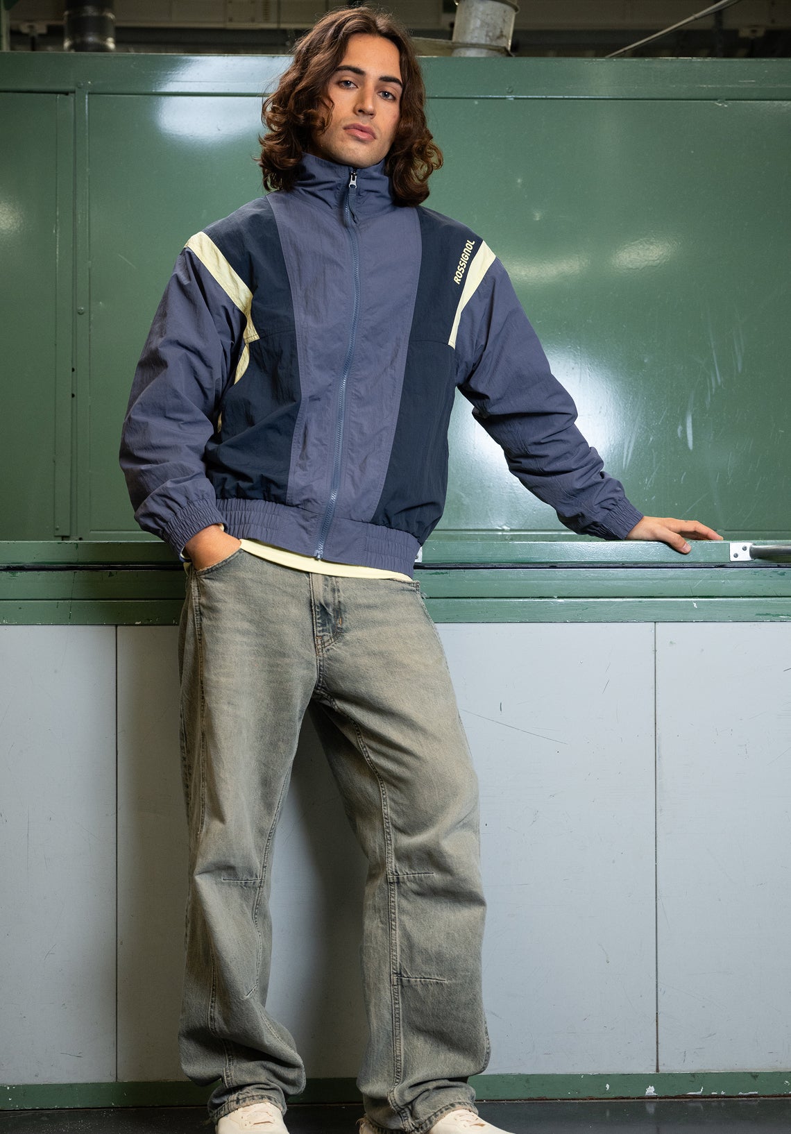 A person standing wears a blue and beige zip-up jacket with a high collar, paired with jeans, in an industrial indoor setting.