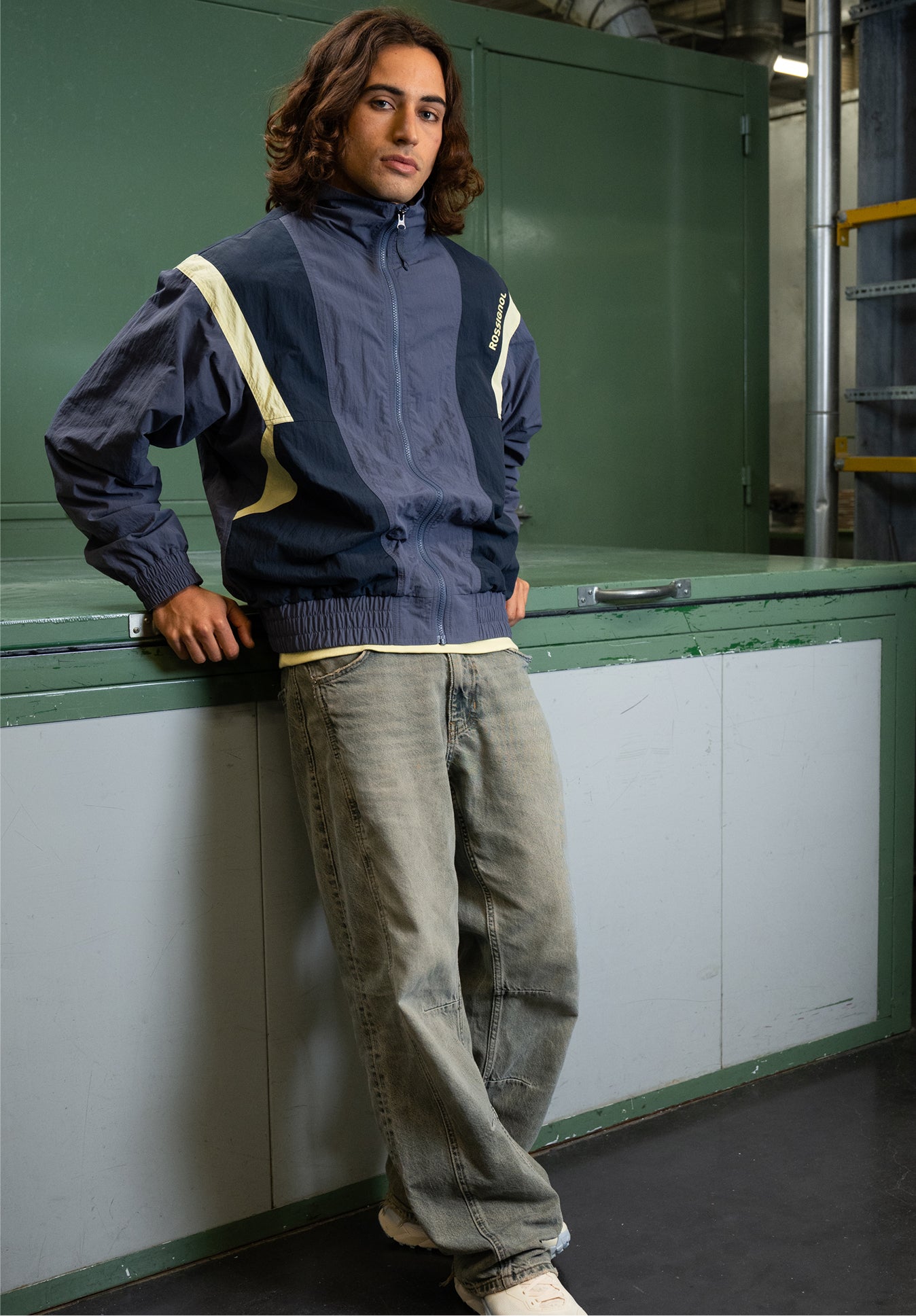 A person leaning against a piece of furniture wears a blue and beige zip-up jacket and jeans, in a workshop-style indoor environment