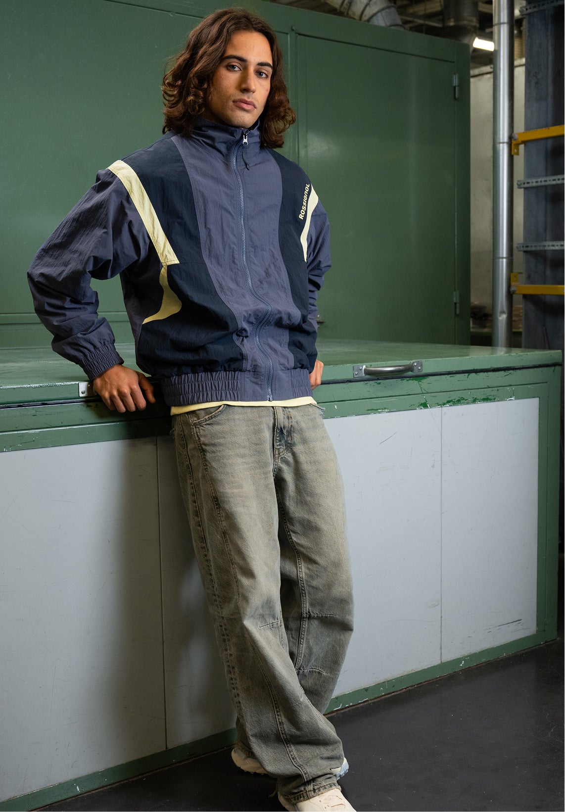A person leaning against a piece of furniture wears a blue and beige zip-up jacket and jeans, in a workshop-style indoor environment.