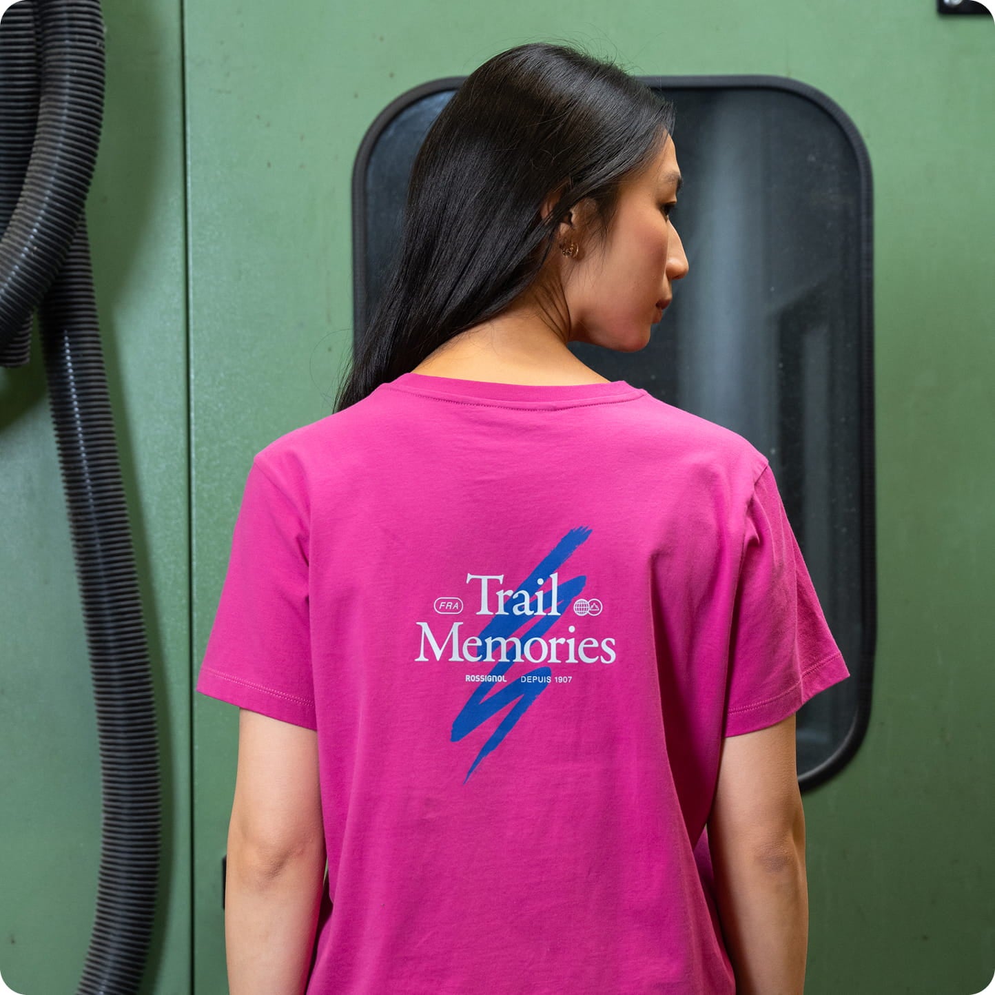 Rear view of a person wearing a pink T-shirt with a graphic design and printed text, in an industrial environment.