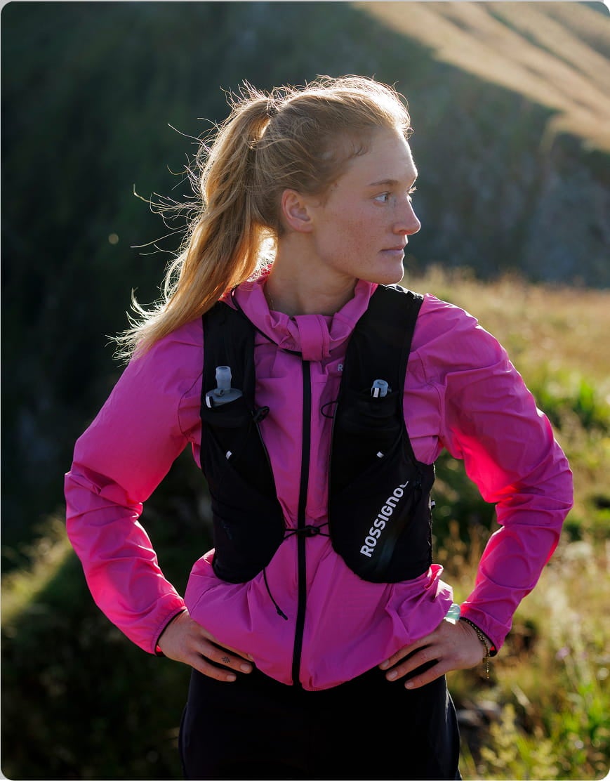 Atleta con giacca da trail rosa e zaino di idratazione in ambiente montano.