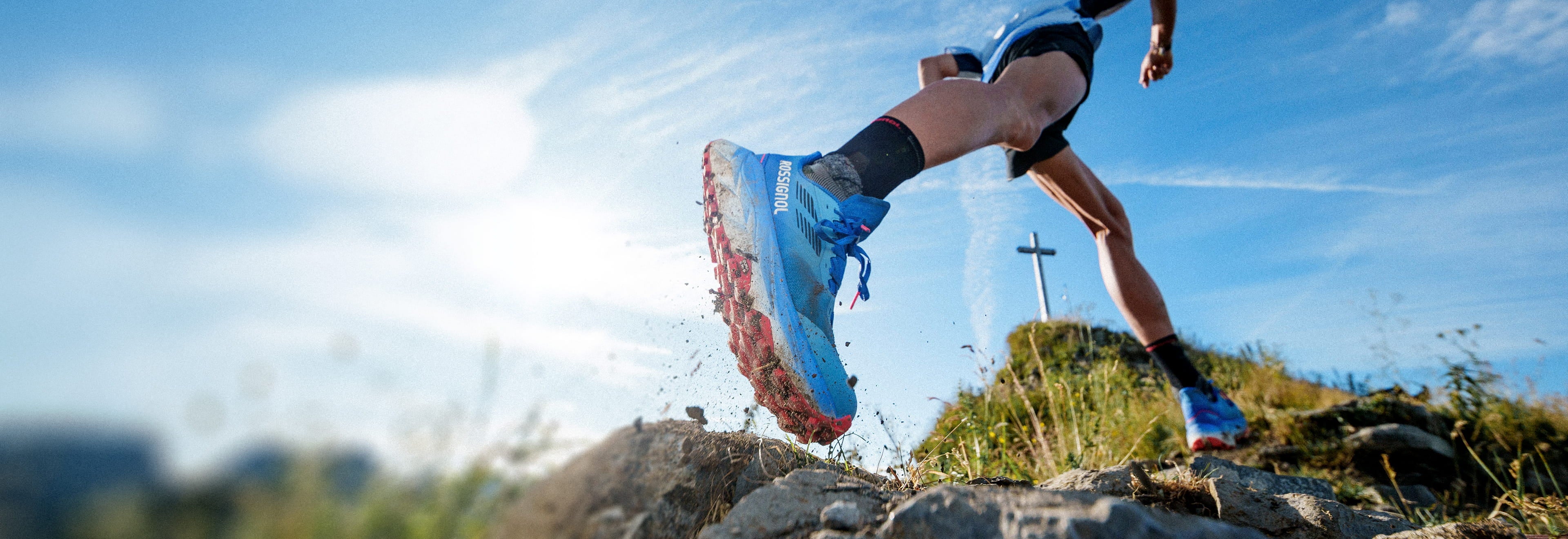 Corridore in salita su un sentiero roccioso con scarpe da trail Rossignol Vercors, con paesaggio montano soleggiato sullo sfondo.
