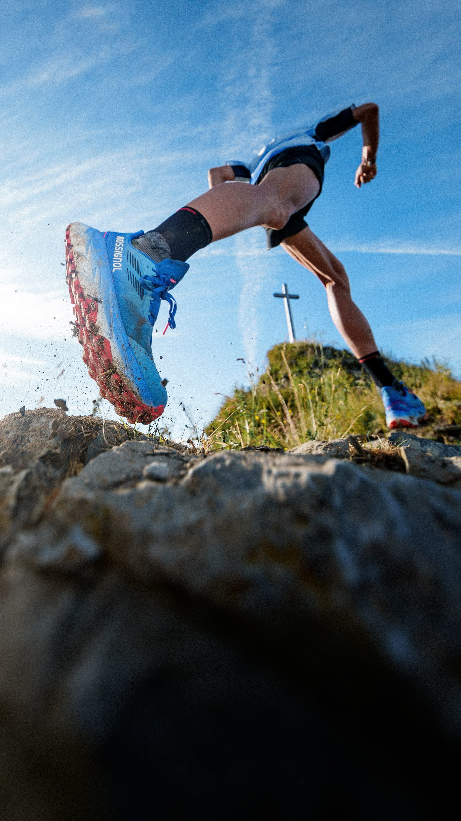 Corridore in salita su un sentiero roccioso con scarpe da trail Rossignol Vercors, con paesaggio montano soleggiato sullo sfondo.