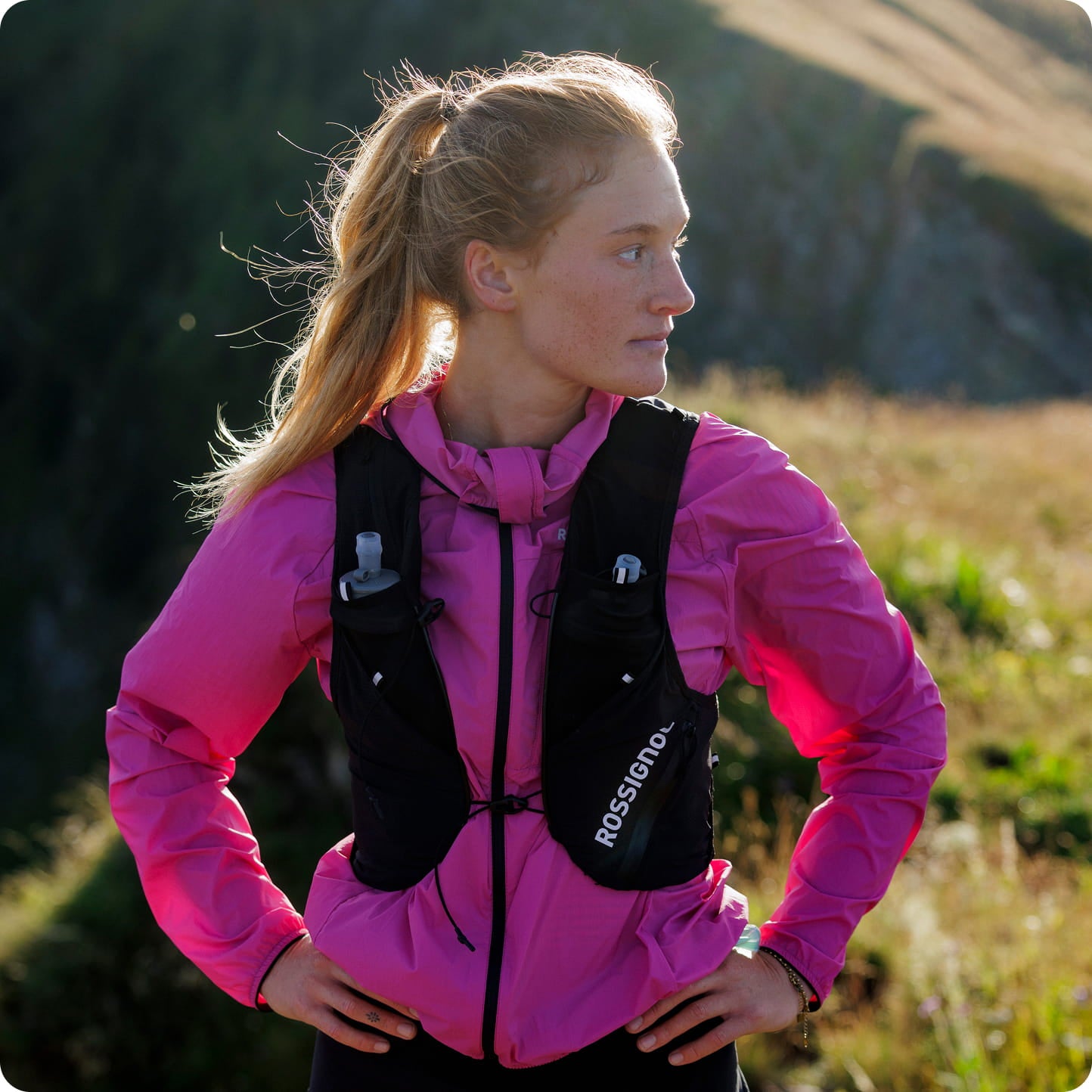 Atleta con giacca da trail rosa e zaino di idratazione in ambiente montano.