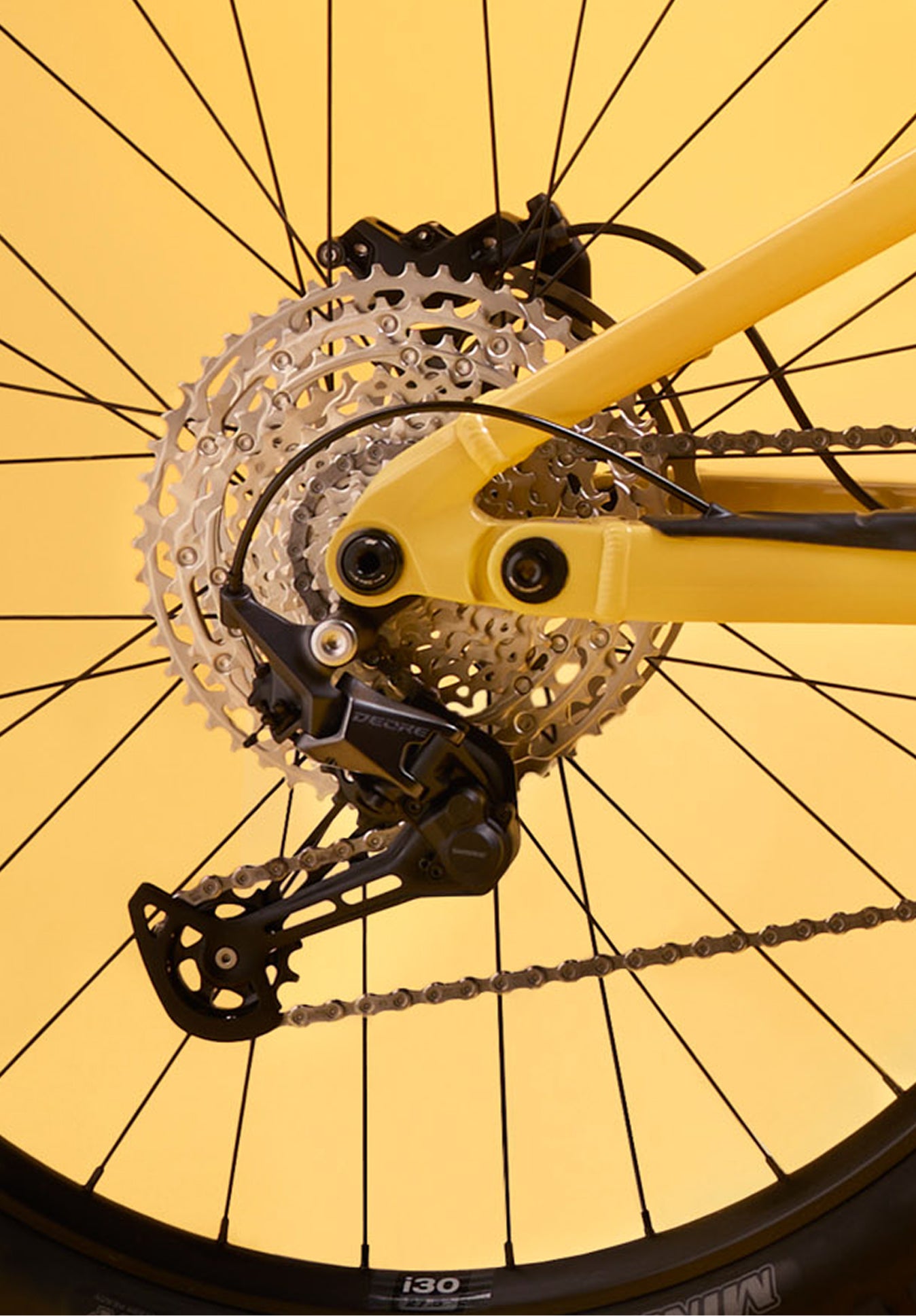 Close-up of the rear drivetrain of the Rossignol After Hours Deore 12, showing cassette, derailleur, and chain on a spoked rear wheel with a yellow frame.