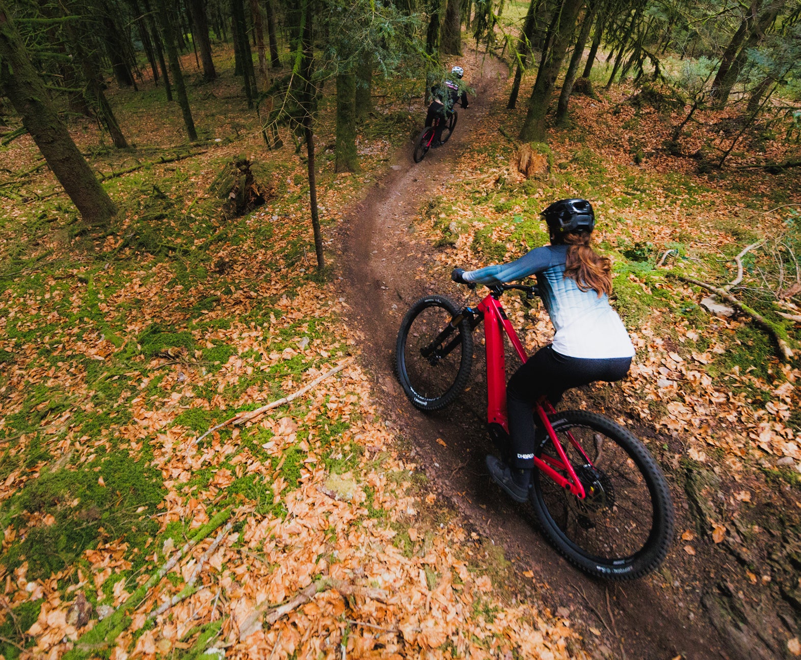 Riders on Rossignol After Hours electric mountain bikes riding along a winding forest trail, seen from above, on a narrow leaf-covered path among trees.