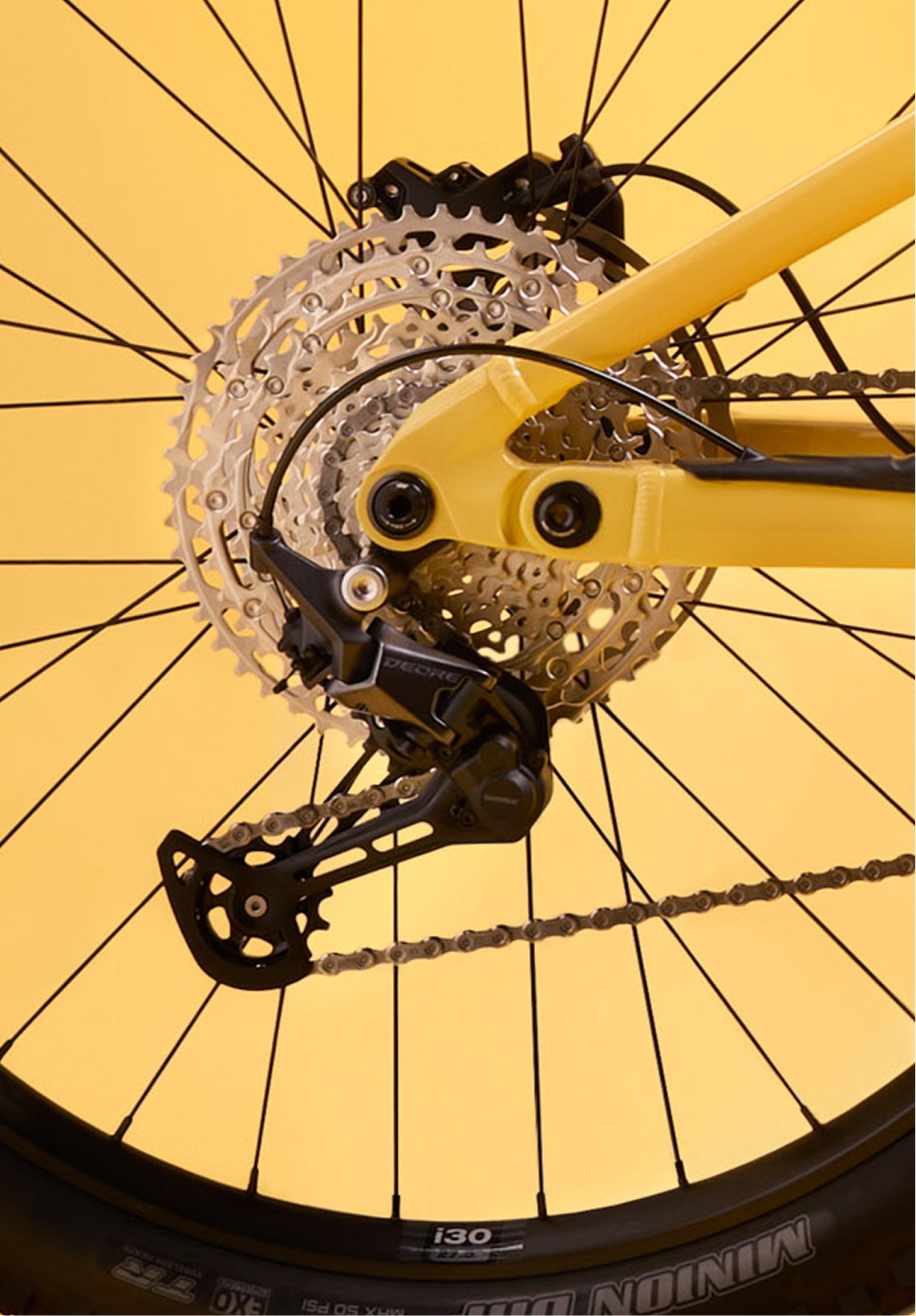 Close-up of the rear drivetrain of the Rossignol After Hours Deore 12, showing cassette, derailleur, and chain on a spoked rear wheel with a yellow frame.