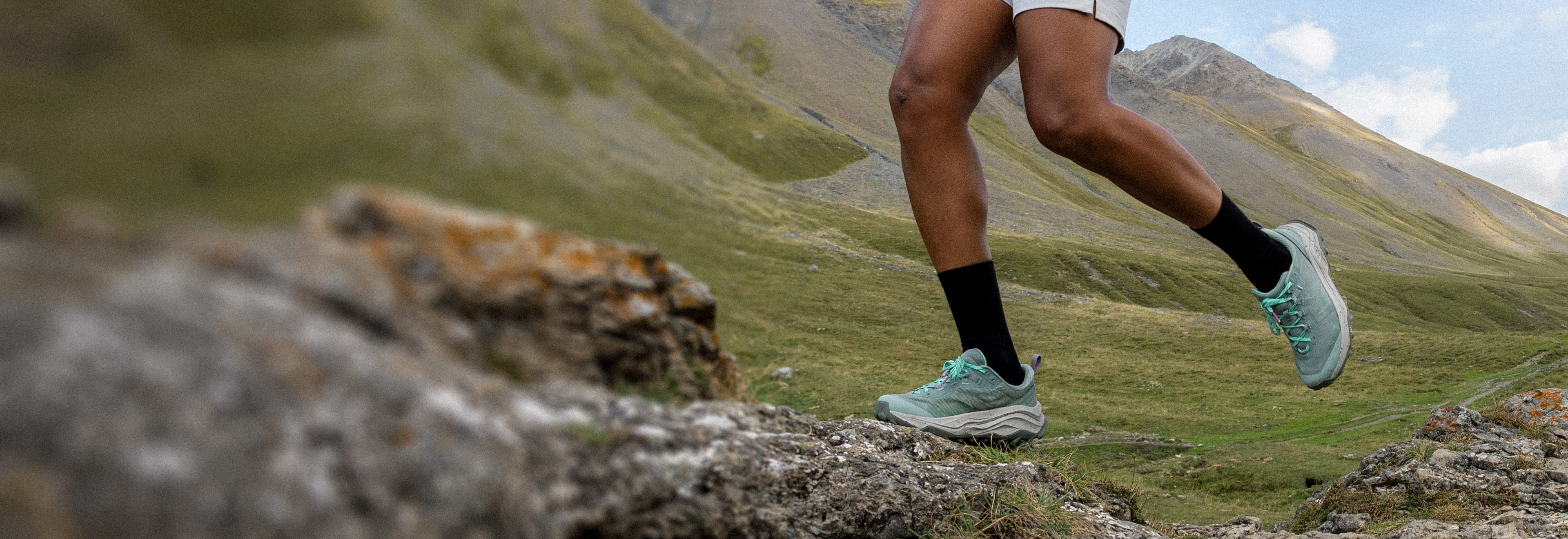 Nahaufnahme der Beine einer Person beim aktiven Wandern in den Bergen. Sie trägt hellgrüne Rossignol-Sneaker, schwarze Socken und ecrufarbene Shorts und läuft auf einem steinigen Pfad vor dem Hintergrund von grünen Tälern und Bergen.