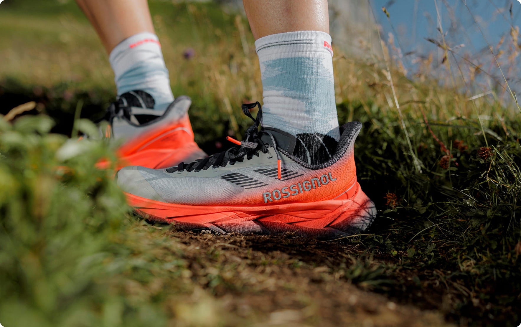 Nahaufnahme einer Person mit Vercors‑Trailrunning-Schuhen auf einem schmalen Erdpfad, umgeben von Gras und Felsen.