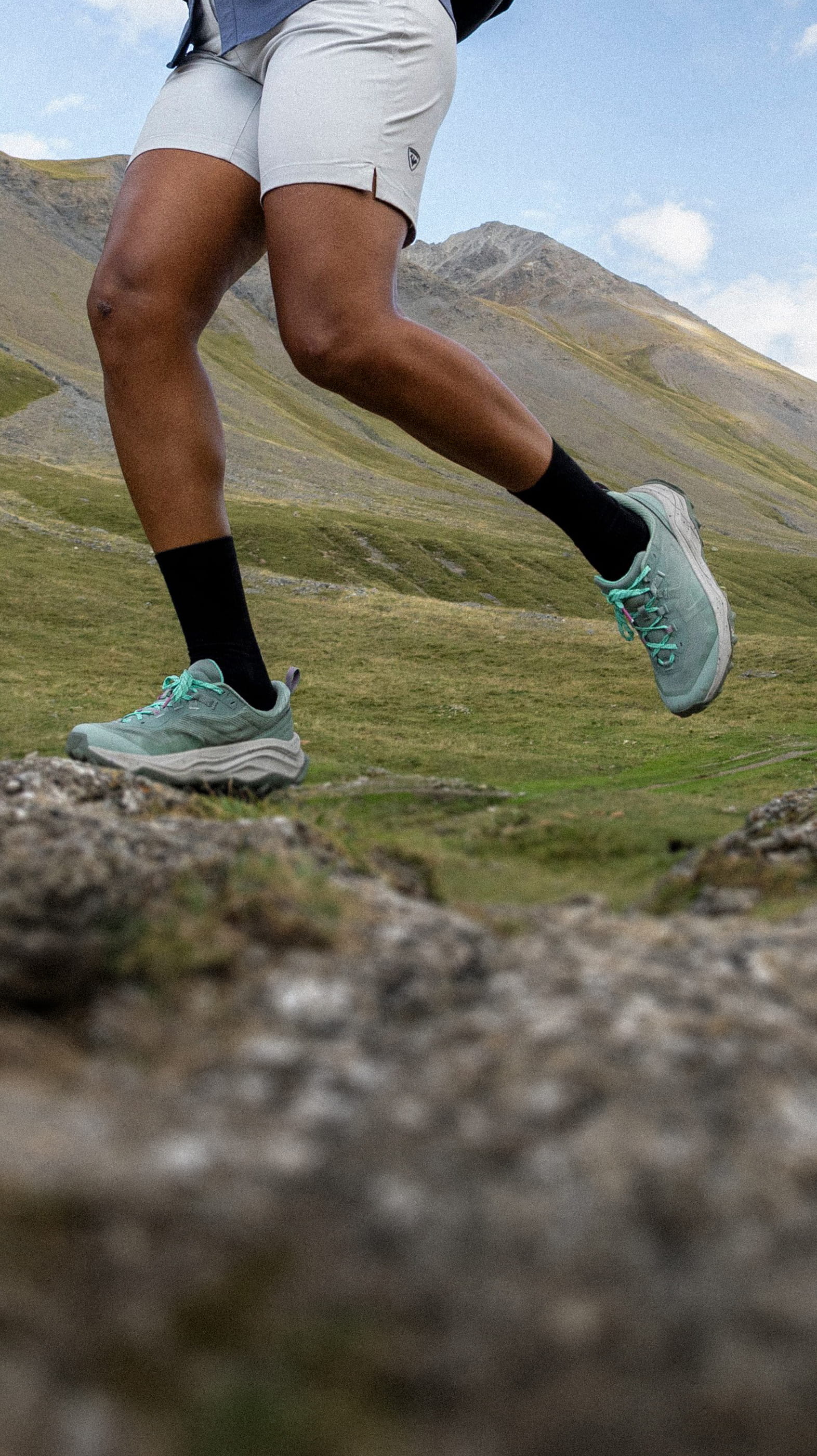 Nahaufnahme der Beine einer Person beim aktiven Wandern in den Bergen. Sie trägt hellgrüne Rossignol-Sneaker, schwarze Socken und ecrufarbene Shorts und läuft auf einem steinigen Pfad vor dem Hintergrund von grünen Tälern und Bergen.