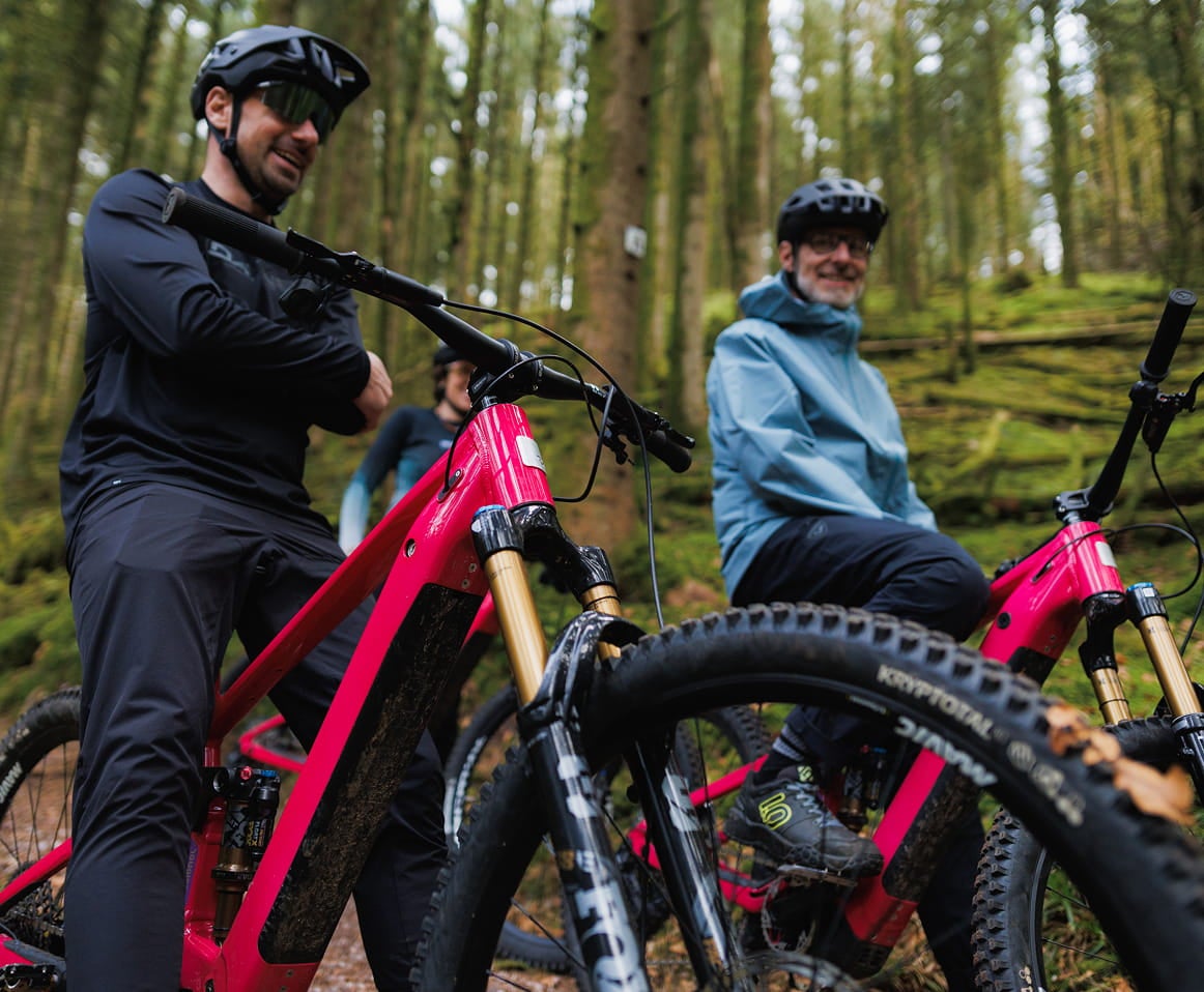 Two athletes, sitting on their Rossignol After Hours bikes. They are chatting during their mountain bike ride in the forest. One is wearing a black outfit and the other a light blue jacket.