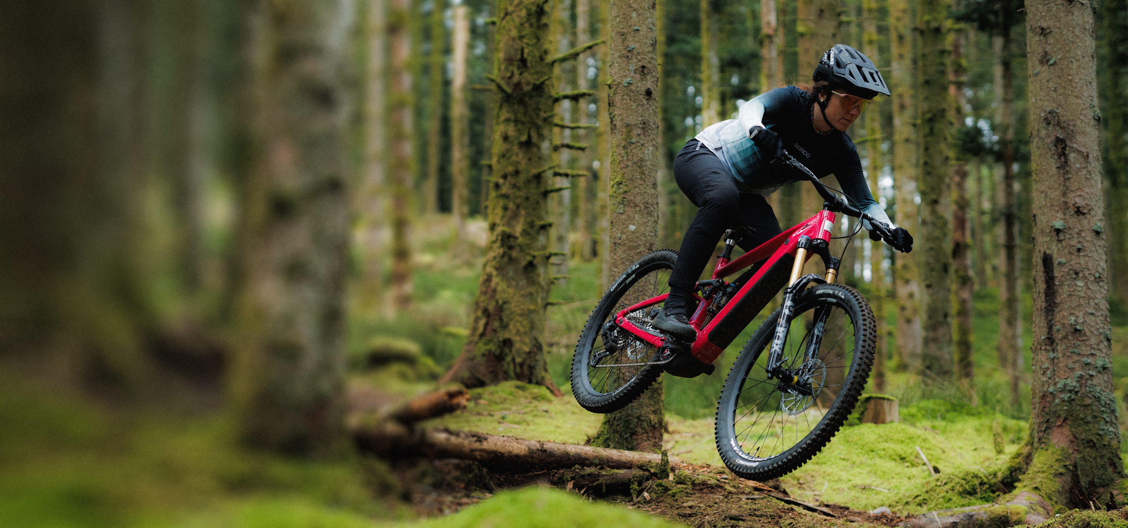 A Rossignol athlete with her After Hours mountain bike riding over a large root in the middle of a lush green forest.