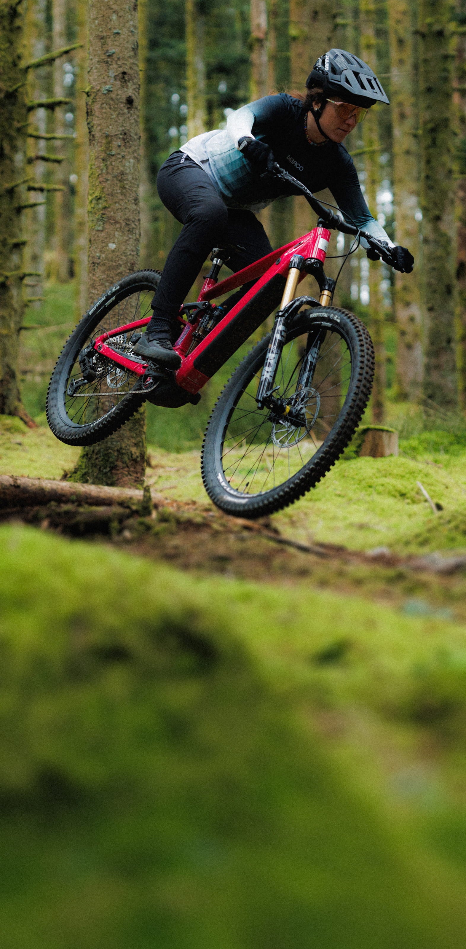 A Rossignol athlete with her After Hours mountain bike riding over a large root in the middle of a lush green forest.
